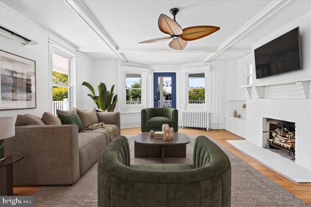 living room featuring beam ceiling, radiator, a brick fireplace, ceiling fan, and wood finished floors