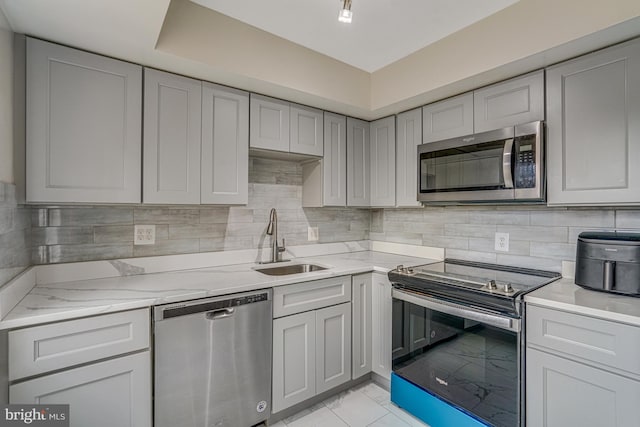 kitchen with gray cabinetry, a sink, appliances with stainless steel finishes, decorative backsplash, and light stone countertops