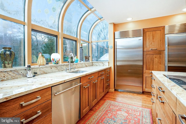 kitchen with light wood finished floors, brown cabinetry, appliances with stainless steel finishes, light stone countertops, and a sink