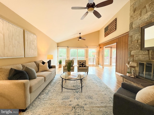 living room featuring a ceiling fan, wood finished floors, baseboards, high vaulted ceiling, and a stone fireplace