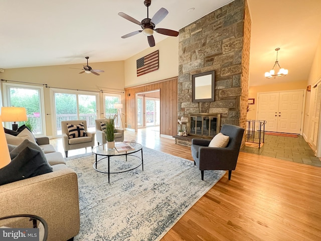 living area featuring a stone fireplace, ceiling fan with notable chandelier, wood finished floors, and high vaulted ceiling