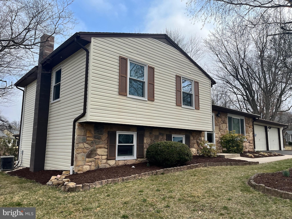 tri-level home featuring stone siding, cooling unit, a front yard, a garage, and a chimney