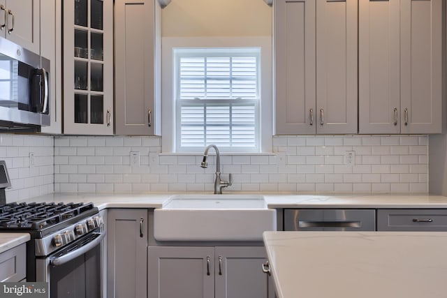 kitchen featuring gray cabinetry, decorative backsplash, a sink, glass insert cabinets, and appliances with stainless steel finishes