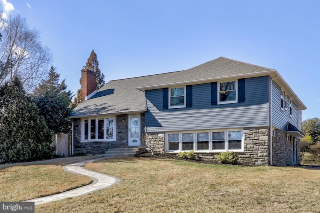 split level home featuring stone siding, roof with shingles, a chimney, and a front yard