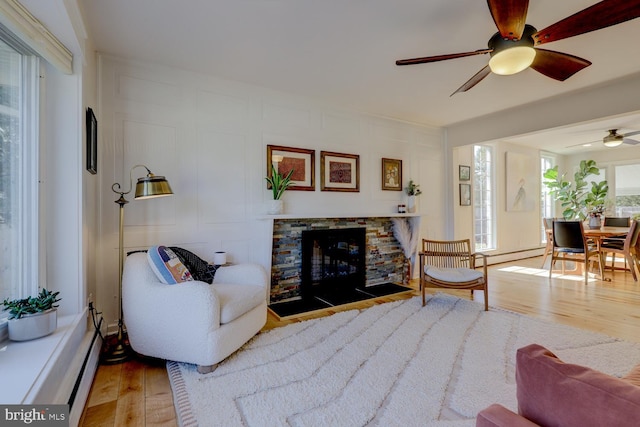 living room with a stone fireplace, a baseboard radiator, a decorative wall, wood finished floors, and a ceiling fan