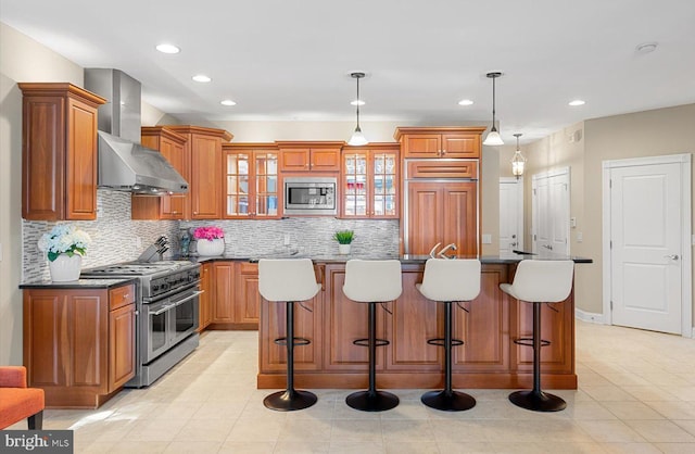 kitchen featuring a breakfast bar, built in appliances, dark countertops, wall chimney exhaust hood, and brown cabinets