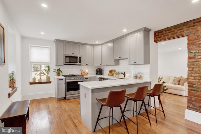 kitchen featuring gray cabinetry, stainless steel appliances, a peninsula, light wood-type flooring, and a kitchen bar