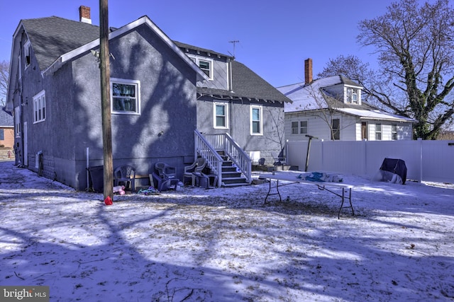 snow covered back of property with a shingled roof, fence, and stucco siding