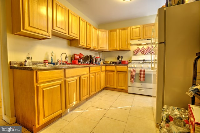 kitchen with white appliances, light tile patterned floors, dark countertops, under cabinet range hood, and a sink