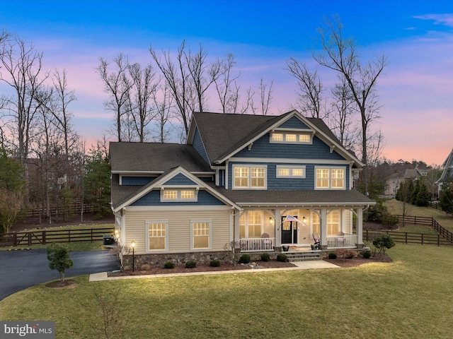 view of front facade featuring aphalt driveway, covered porch, fence, stone siding, and a lawn