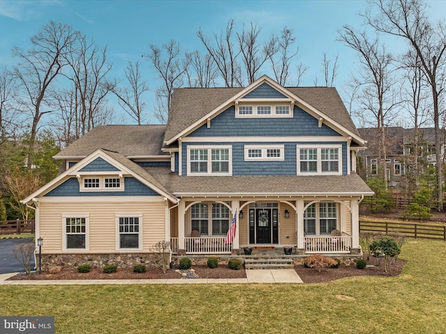 view of front facade with stone siding, roof with shingles, covered porch, fence, and a front lawn