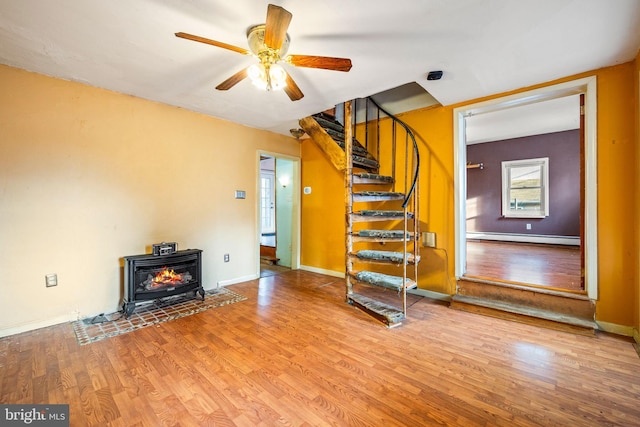 unfurnished living room featuring ceiling fan, stairway, wood finished floors, a wood stove, and a baseboard heating unit