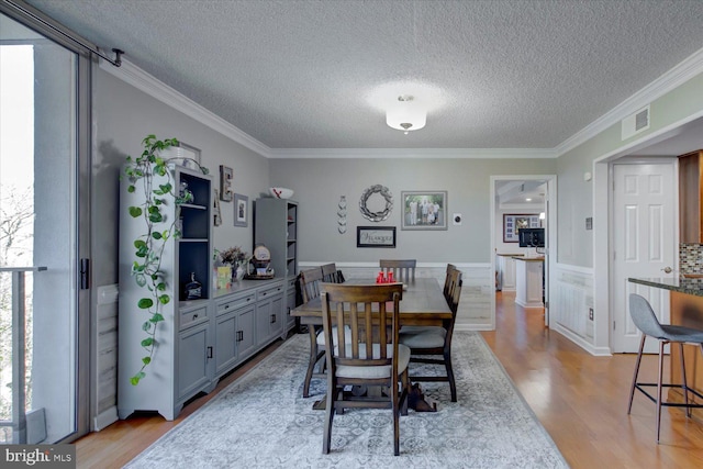 dining space with light wood finished floors, visible vents, a wainscoted wall, ornamental molding, and a textured ceiling