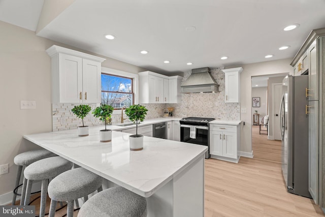 kitchen with light stone counters, premium range hood, light wood finished floors, a peninsula, and appliances with stainless steel finishes