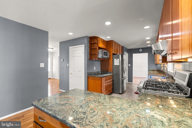 kitchen with dark stone countertops, range hood, stainless steel appliances, open shelves, and a sink