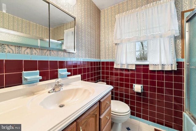 bathroom with tile walls, wainscoting, toilet, and wallpapered walls