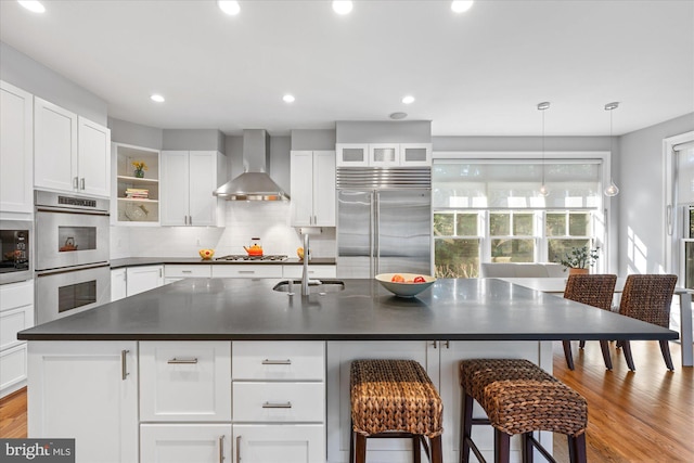 kitchen with a sink, open shelves, dark countertops, wall chimney range hood, and built in appliances