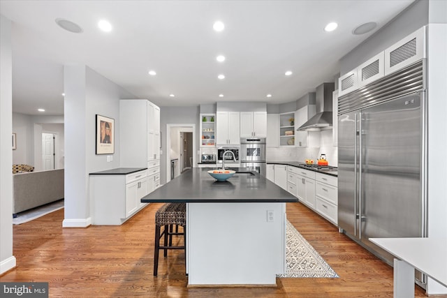 kitchen with open shelves, light wood-style floors, appliances with stainless steel finishes, dark countertops, and wall chimney exhaust hood