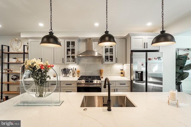 kitchen featuring wall chimney exhaust hood, glass insert cabinets, light stone countertops, stainless steel appliances, and a sink