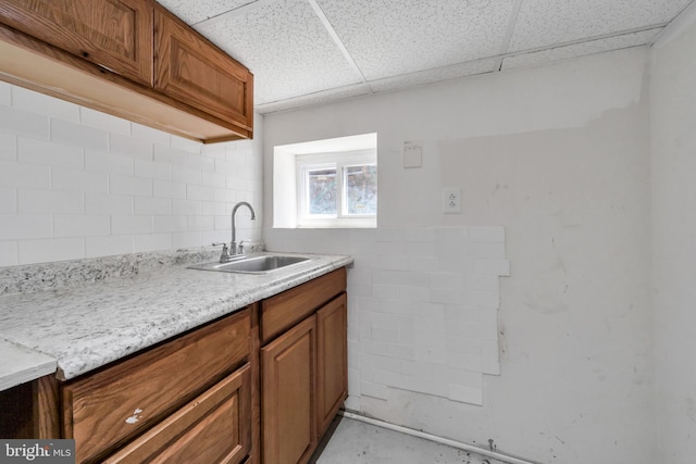kitchen with a sink, a paneled ceiling, brown cabinetry, backsplash, and concrete flooring