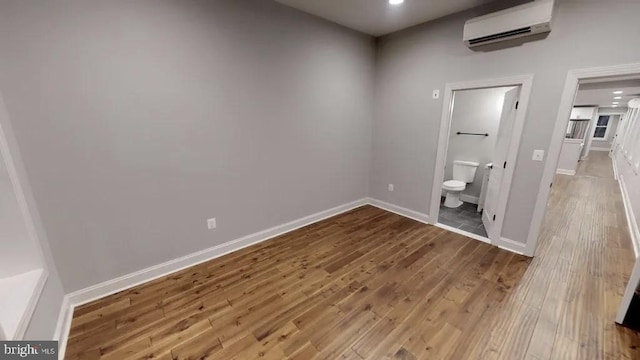 unfurnished bedroom with an AC wall unit, ensuite bath, wood finished floors, and baseboards