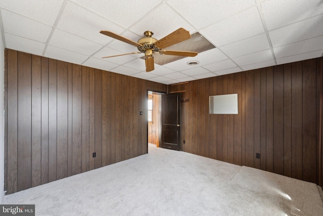 carpeted empty room with wood walls, ceiling fan, visible vents, and a drop ceiling