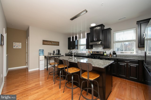 kitchen with backsplash, under cabinet range hood, stainless steel range with gas stovetop, a center island with sink, and a sink