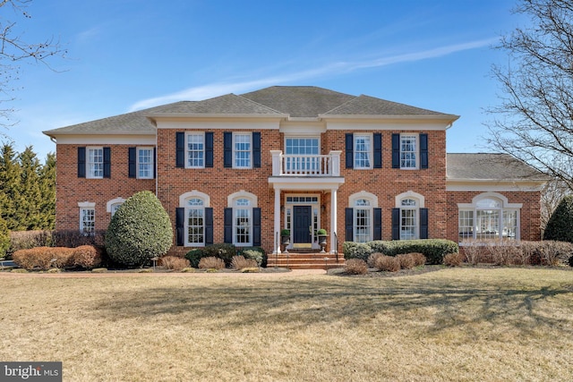 colonial-style house featuring roof with shingles, brick siding, a front lawn, and a balcony