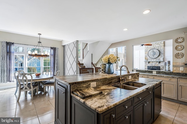 kitchen with light tile patterned floors, a sink, dark stone countertops, an island with sink, and dishwashing machine