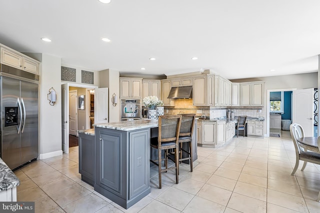 kitchen featuring light tile patterned floors, backsplash, cream cabinets, stainless steel built in refrigerator, and under cabinet range hood