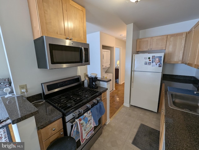 kitchen with dark stone counters, stainless steel appliances, stacked washing maching and dryer, light brown cabinets, and a sink