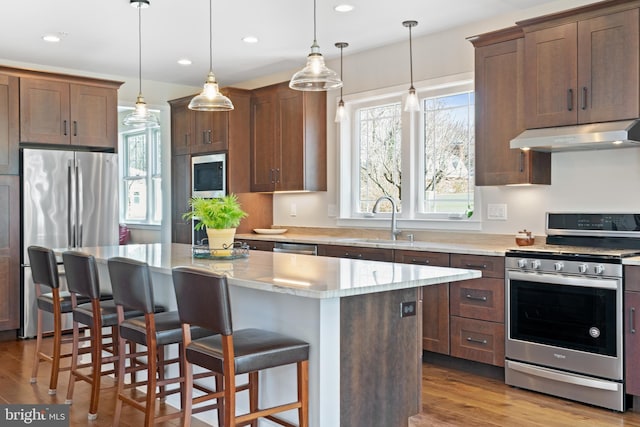 kitchen with a kitchen island, under cabinet range hood, appliances with stainless steel finishes, wood finished floors, and a sink