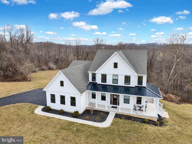 back of property with a yard, a shingled roof, a porch, a standing seam roof, and a view of trees