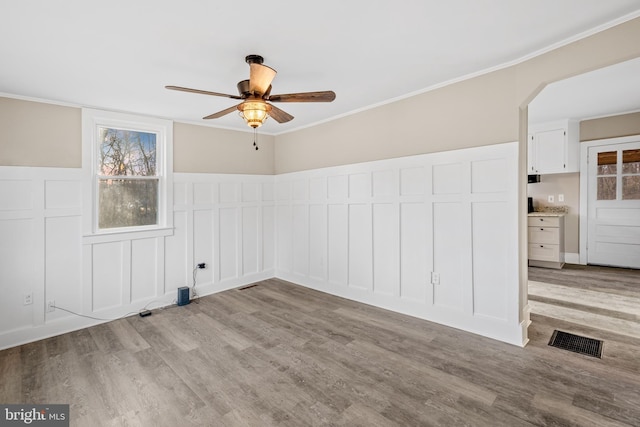 unfurnished room featuring wood finished floors, a wainscoted wall, visible vents, ceiling fan, and crown molding