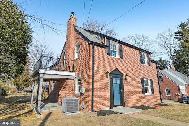 view of front facade featuring a balcony, central air condition unit, a chimney, and brick siding