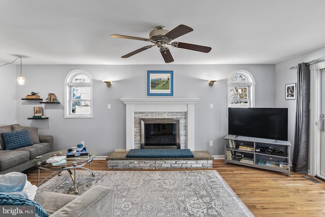 living room with a brick fireplace, ceiling fan, baseboards, and wood finished floors