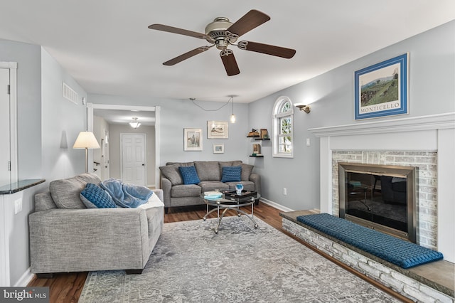 living area featuring dark wood finished floors, visible vents, a ceiling fan, a brick fireplace, and baseboards