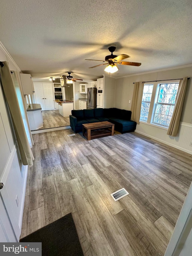 unfurnished living room featuring a textured ceiling, wood finished floors, visible vents, a ceiling fan, and ornamental molding