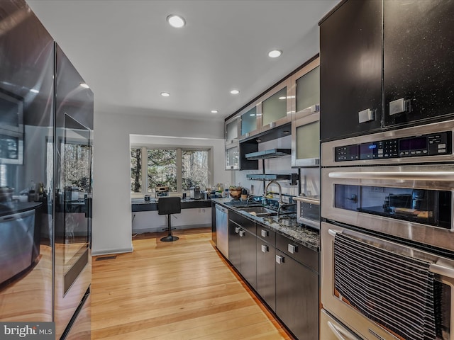 kitchen featuring light wood finished floors, stainless steel appliances, glass insert cabinets, a sink, and dark stone counters