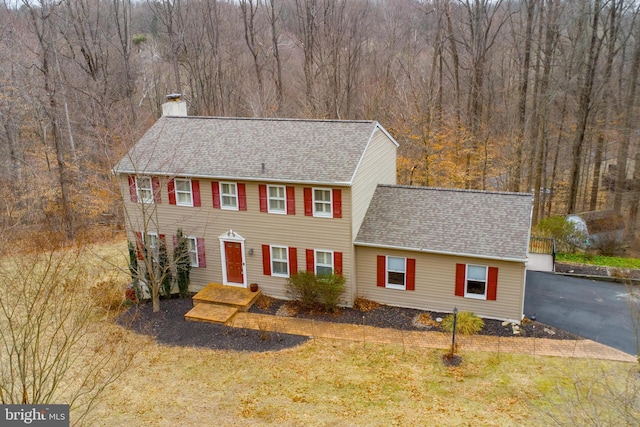 colonial house featuring a front lawn, a wooded view, a chimney, and roof with shingles