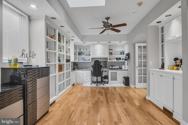 office area with a skylight, arched walkways, built in study area, ceiling fan, and light wood-type flooring