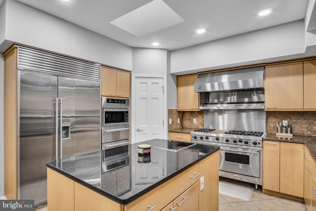 kitchen with light tile patterned floors, premium appliances, dark stone countertops, light brown cabinetry, and wall chimney range hood