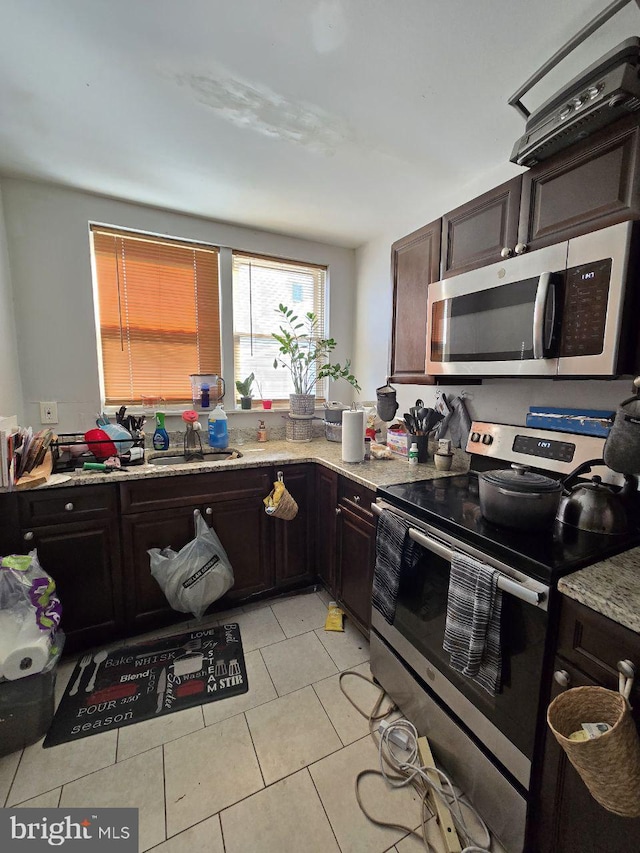 kitchen with light tile patterned floors, stainless steel appliances, a sink, dark brown cabinets, and light stone countertops