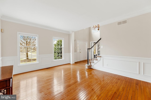 unfurnished living room featuring visible vents, stairs, light wood-style floors, wainscoting, and crown molding