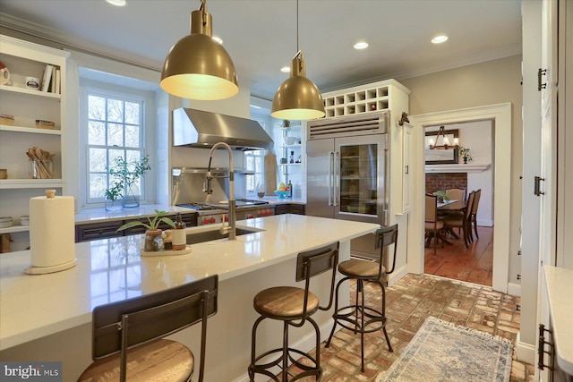 kitchen with brick floor, recessed lighting, built in refrigerator, ornamental molding, and wall chimney range hood