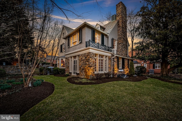property exterior at dusk with stone siding, stucco siding, a lawn, and a balcony