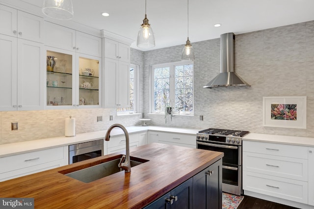 kitchen with butcher block counters, a sink, white cabinetry, wall chimney range hood, and appliances with stainless steel finishes