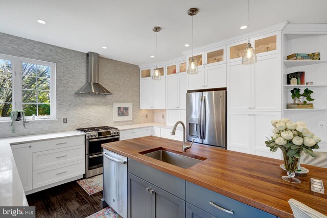 kitchen featuring wall chimney exhaust hood, butcher block counters, stainless steel appliances, pendant lighting, and a sink