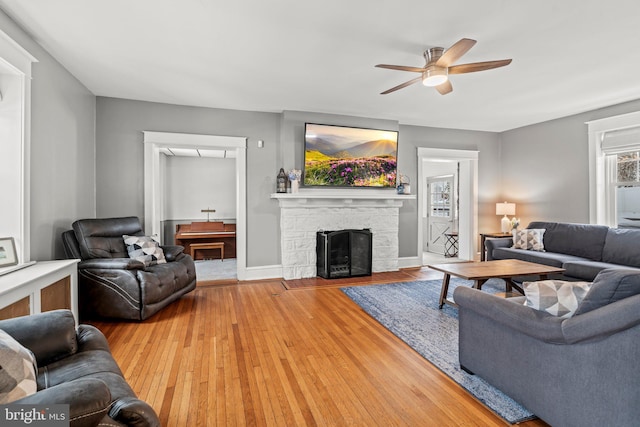 living area with a ceiling fan, baseboards, hardwood / wood-style floors, and a stone fireplace