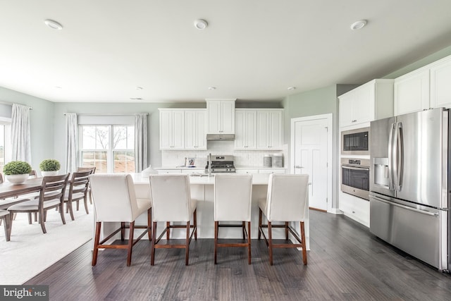 kitchen featuring a center island with sink, stainless steel appliances, light countertops, under cabinet range hood, and backsplash